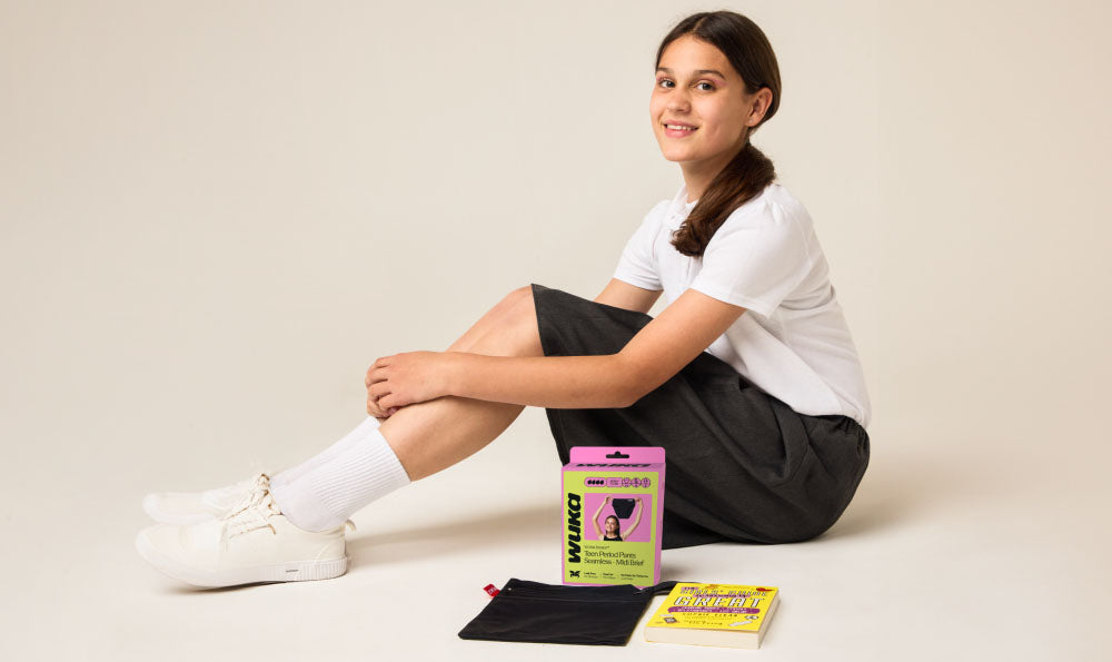 Teen girl in school uniform sitting beside WUKA period pants, storage bag, and puberty education booklet – supporting WUKA’s mission to empower teens with accessible, period-positive puberty education