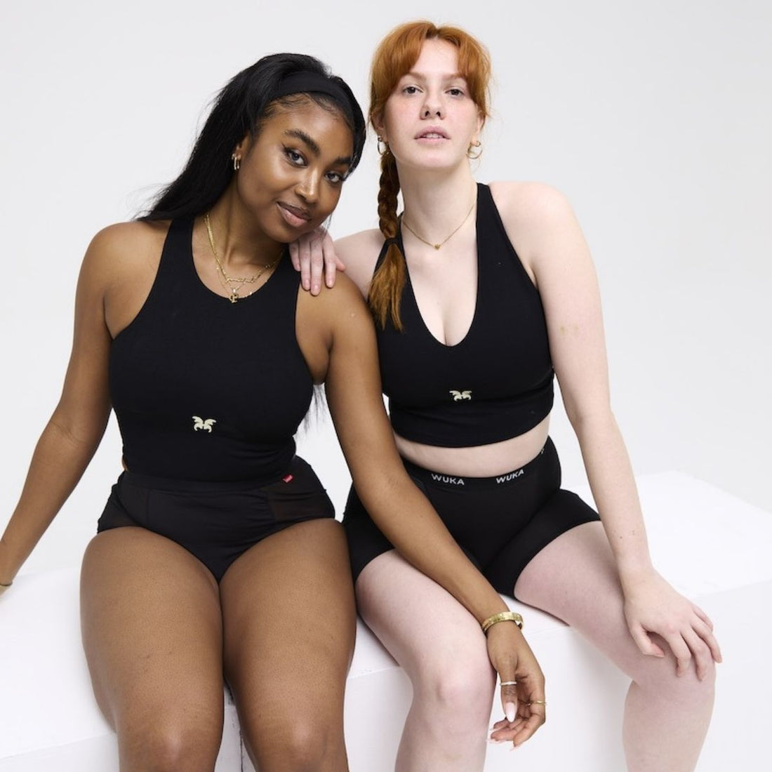 Two young women wearing black WUKA period underwear and matching tops, seated confidently against a white studio background, arms linked in solidarity.