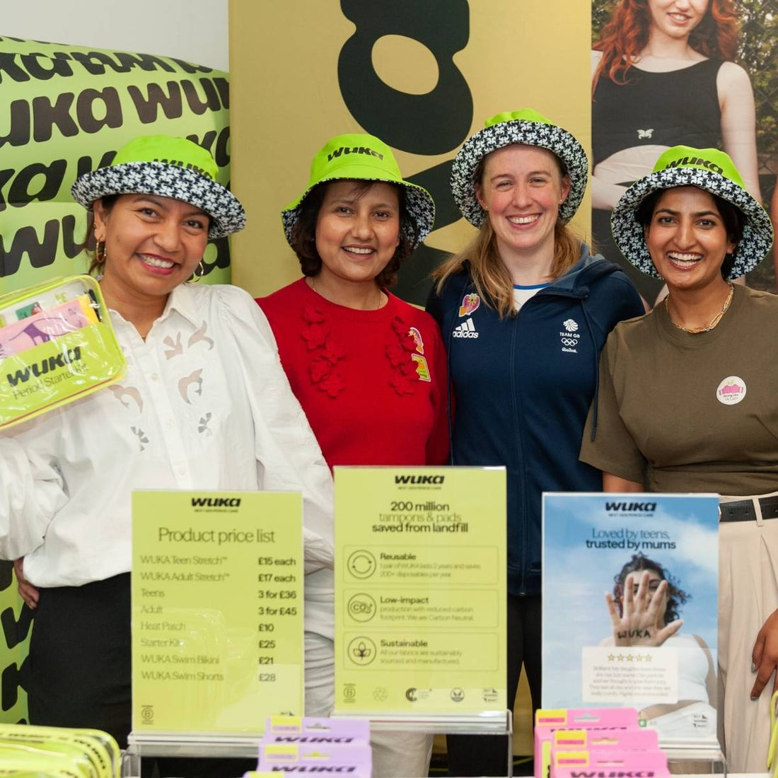 Ruby Raut and the WUKA team smile at a pop-up event, wearing branded hats and holding period kits, celebrating reusables and community impact with pride.