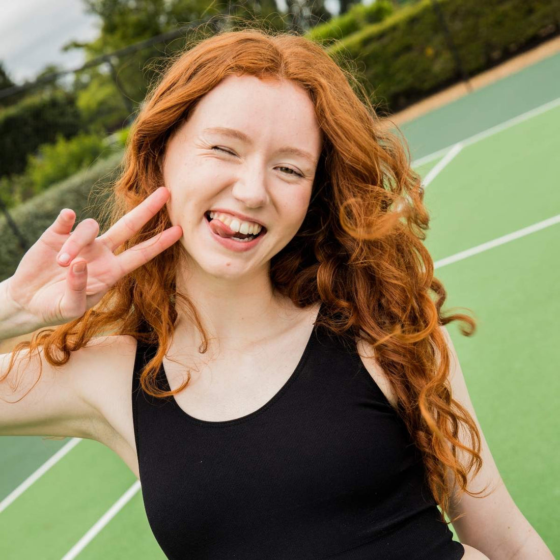 Teen girl with long red curly hair smiles playfully on a tennis court, sticking out her tongue and flashing a peace sign while wearing a black tank top.