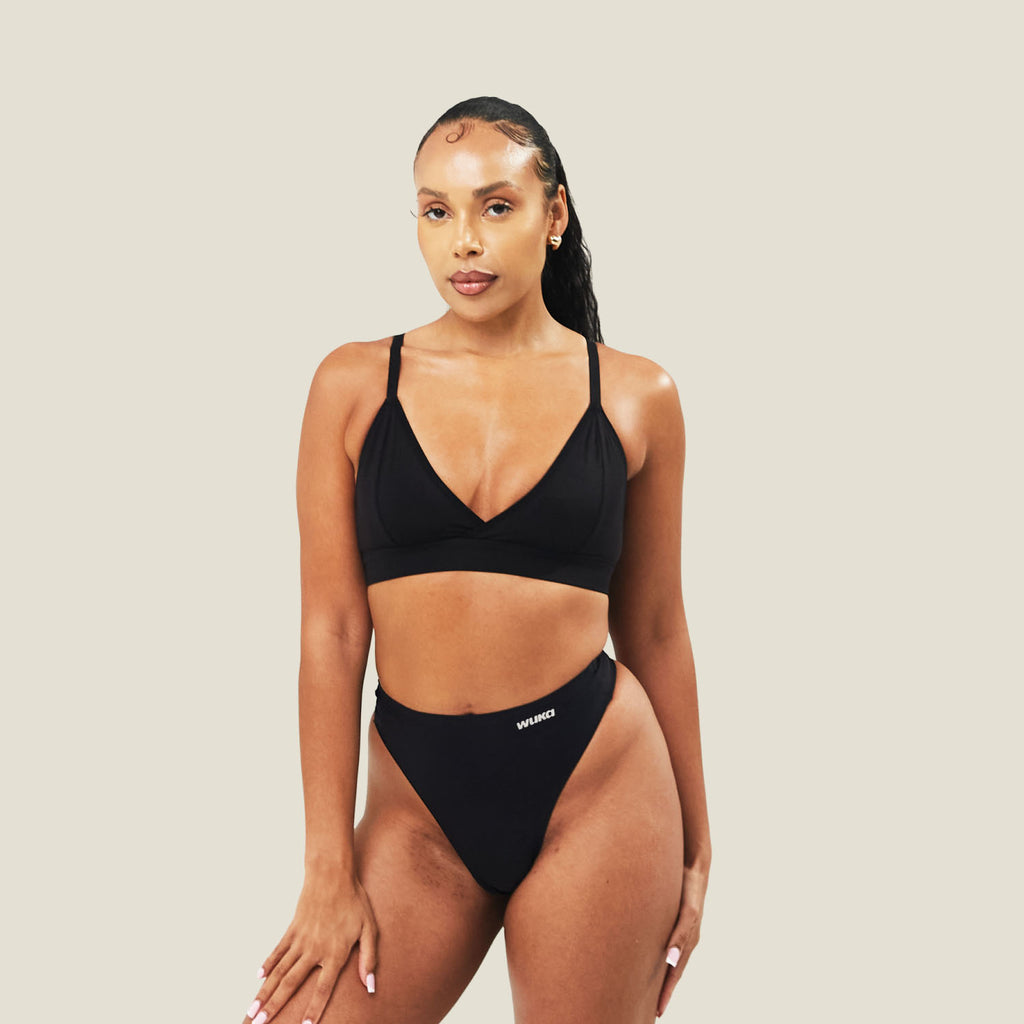 Model wearing the WUKA Stretch Seamless Mid Period Thong for light flow in black, with a matching black bralette, standing against a cream background.