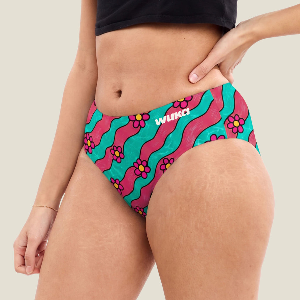 A close-up of a model wearing teen midi brief period pants with a vibrant retro pattern of pink and teal wavy stripes and magenta flowers. The brand name 'WUKA' is printed in white on the front right hip.