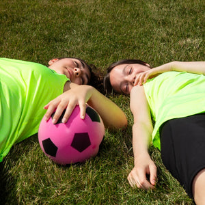 Two girls lying on grass with a football, representing rest and recovery during sport while managing periods with confidence